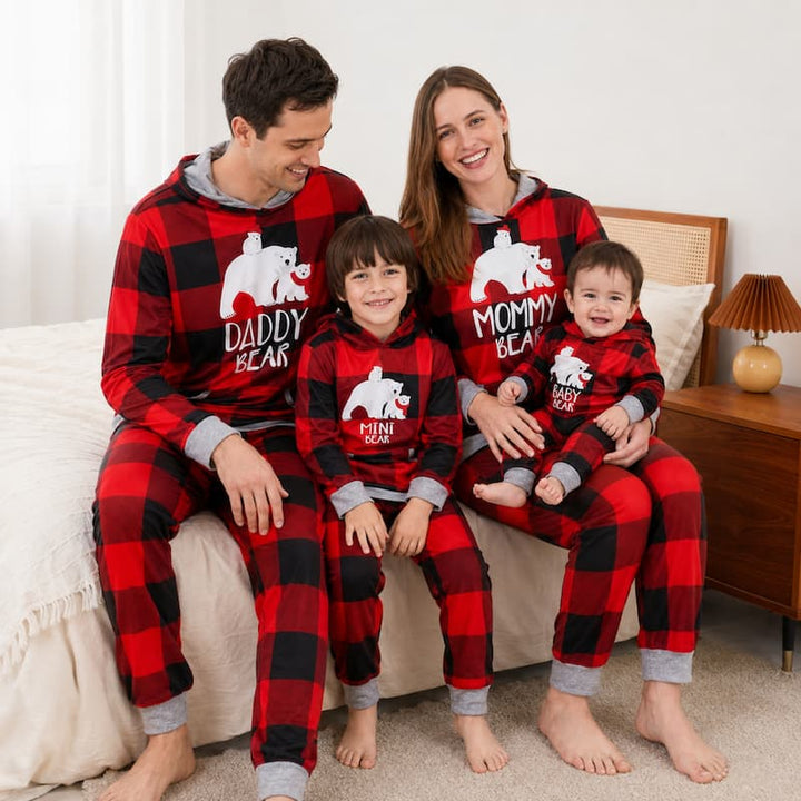 A family of four wearing matching red and black buffalo plaid hooded pajamas with white bear graphics and text, sitting together on a bed in a bedroom setting.