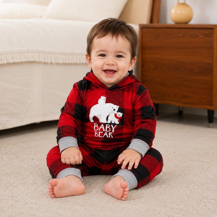 A smiling baby sits on a beige carpet wearing a red and black buffalo plaid hooded onesie with gray cuffs and a white 'BABY BEAR' graphic on the chest. The background shows a cozy bedroom setting with a bed and wooden nightstand.