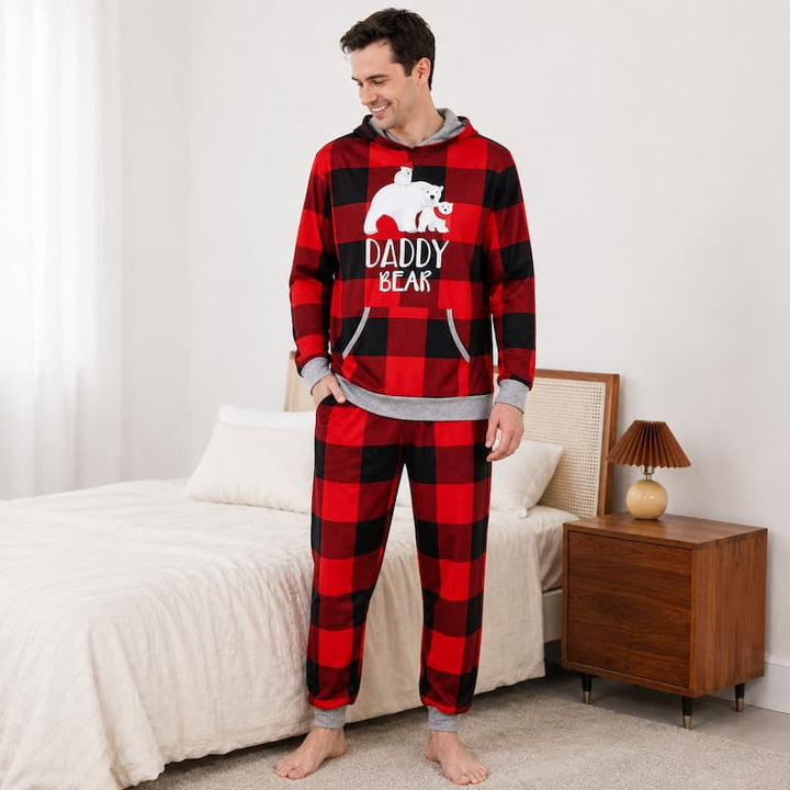 A man stands barefoot in a bedroom wearing a red and black buffalo plaid hooded sweatshirt and matching sweatpants with a white bear graphic and the words 'DADDY BEAR' on the front. The room includes a bed, nightstand, lamp, and light-colored walls.