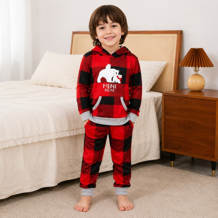 A young boy stands barefoot in a bedroom wearing a red and black buffalo plaid hooded pajama set with a white polar bear graphic and the words 'MINI BEAR' on the front. The bedroom has a beige bed, a wooden nightstand with a lamp, and a light-colored wall.