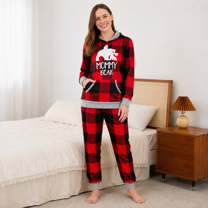 A woman stands barefoot in a bedroom wearing a red and black buffalo plaid pajama set with a gray hood and cuffs. The hoodie features a white polar bear graphic and the text 'MOMMY BEAR'.