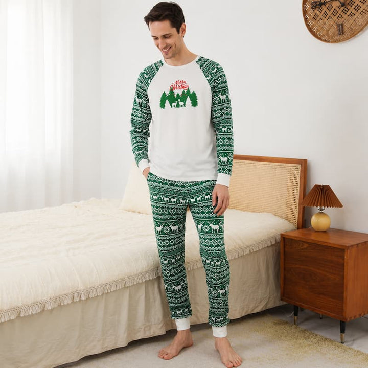 A man stands barefoot in a bedroom wearing a holiday-themed long-sleeve pajama set. The set has a white crewneck top with green and red Christmas tree graphics on the chest, green raglan sleeves, and matching green patterned pants with white cuffs.