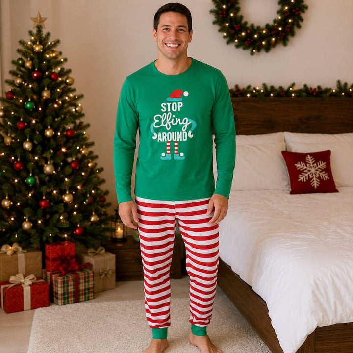 A man stands in a bedroom wearing a green long-sleeve Christmas shirt with the text 'STOP Elfing AROUND' and red-and-white striped pajama pants. A decorated Christmas tree, bed, and wreath are visible in the background.